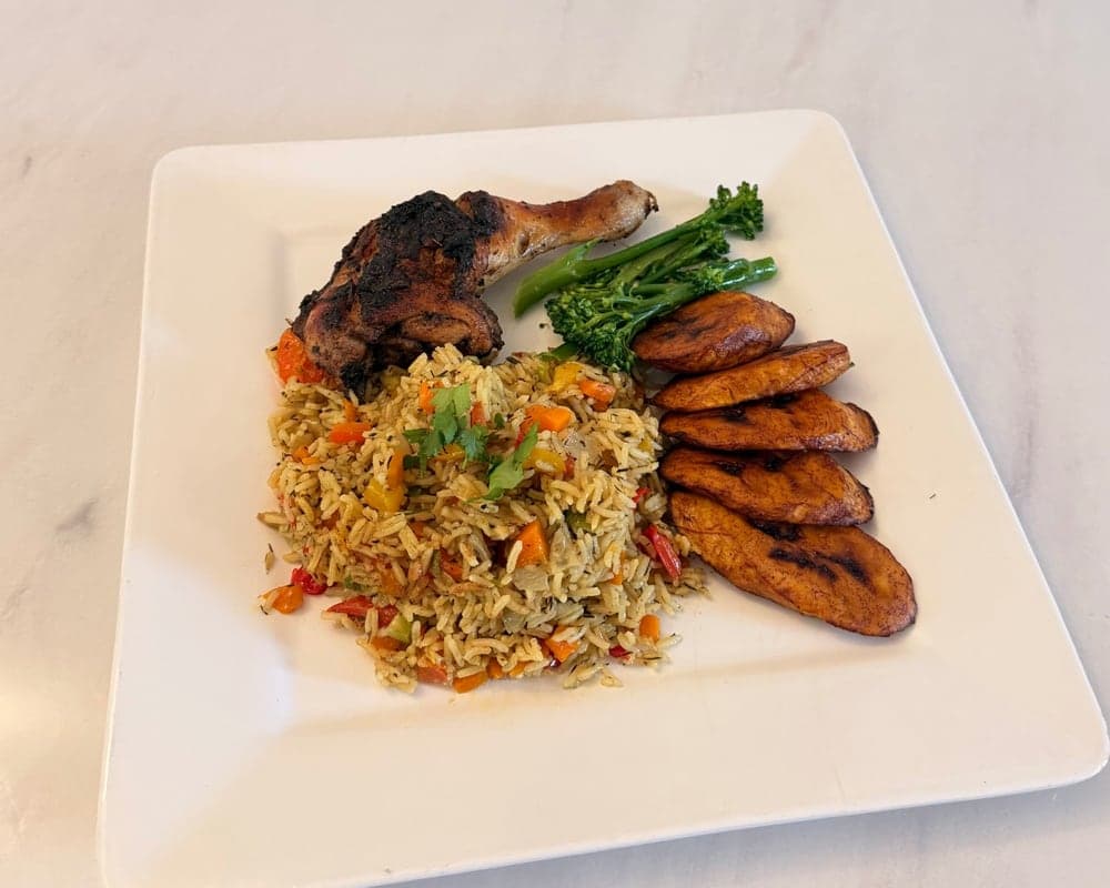A square white plate of Nigerian fried rice with grilled chicken, steamed tenderstem broccoli and fried plantain.