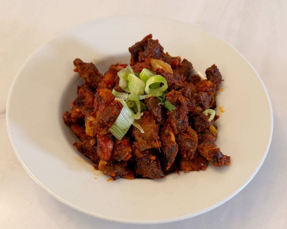 A white bowl of gizdodo, chicken gizzards and fried plantain in a red pepper sauce, topped with sliced spring onion.