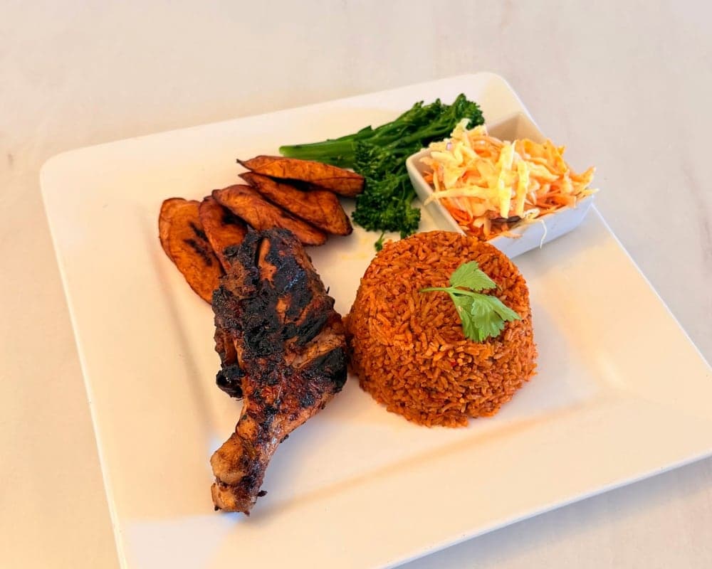 A square white plate of party jollof rice with grilled chicken, coleslaw, steamed tenderstem broccoli and fried plantain.
