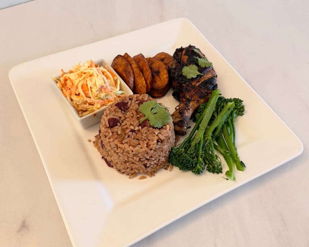 A square white plate of rice and peas with grilled chicken, coleslaw, steamed tenderstem broccoli and fried plantain.